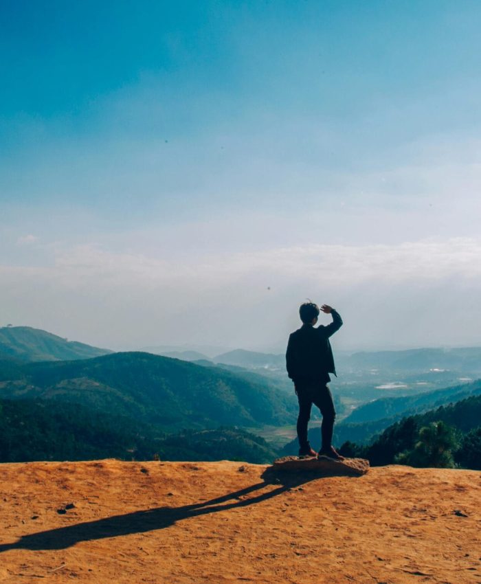 A lone traveler stands on a hilltop, gazing over a vast mountain landscape under a clear blue sky.